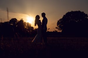 A bride and groom stand close together, gazing at each other in a field at sunset, their silhouettes framed by the warm glow of the sun and trees in the background.