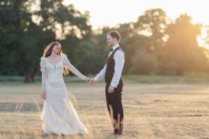 A bride and groom hold hands and smile at each other while standing in a sunlit field, dressed in wedding attire with trees and warm sunlight in the background.