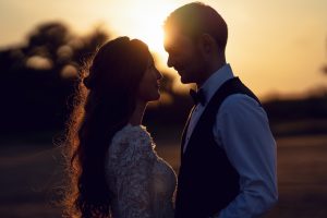 A bride and groom stand close together outdoors at sunset, facing each other and smiling. The sun creates a warm, glowing silhouette around them, highlighting the bride’s long hair and lace dress.