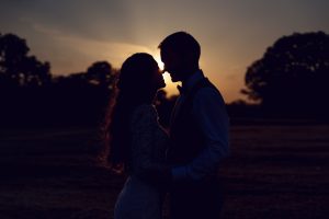 A silhouetted couple stands close together at sunset in an open field, facing each other, with trees in the background and the sun glowing between them.