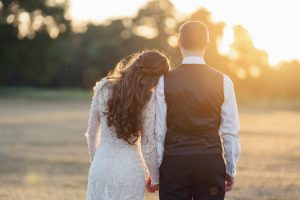 A couple stands outdoors at sunset, facing away. The woman in a white lace dress leans her head on the man's shoulder; he wears a black waistcoat and white shirt. They hold hands in a sunlit field, surrounded by blurred trees.