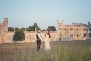 A couple in wedding attire hold hands and smile at the camera, standing in a grassy field with a large, historic stone building in the background under a clear blue sky.