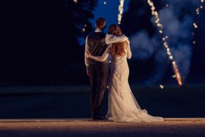 A bride and groom stand arm in arm, facing away, watching fireworks light up the night sky on their wedding day. The bride wears a long white dress, and the groom is in a waistcoat and dress shirt.