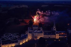Aerial view of a large, illuminated castle at dusk with colourful fireworks bursting in the gardens behind it, surrounded by dark trees and expansive grounds.