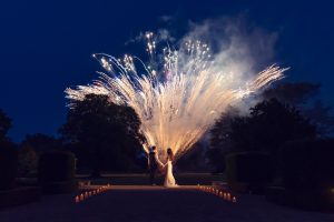 A bride and groom stand hand in hand at night, framed by a dramatic burst of fireworks lighting up the sky behind them, with candles lining the path where they stand.