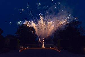A couple stands hand in hand outdoors at night, surrounded by candlelit pathways, whilst bright fireworks burst in the sky above them, creating a dramatic and romantic scene.