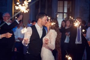 A bride and groom kiss while holding sparklers, surrounded by guests celebrating with sparklers in an evening wedding setting.