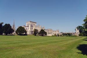 A large historic castle with multiple towers and battlements sits surrounded by lush green lawns and trees under a clear blue sky.
