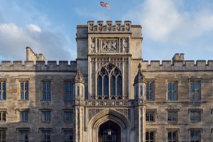 A historic stone building with Gothic architectural details, arched windows, and battlements. An ornate entrance is centred below a carved crest, and a British flag flies above the roof against a partly cloudy sky.