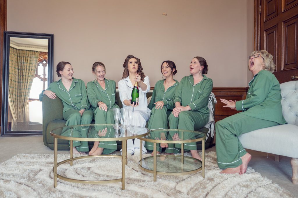 Six women sit on a sofa and chairs in a lounge, all wearing matching green pyjamas except one in white. They laugh and smile as the woman in white opens a bottle of champagne.
