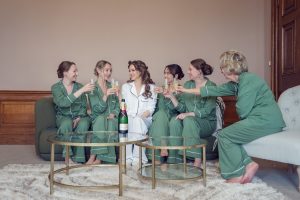 Six women, five in matching green pyjamas and one in white pyjamas, sit together on a sofa and chairs, smiling and raising glasses in a toast, with a bottle on a glass table in front of them.