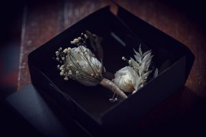 A white buttonhole with small flowers and greenery lies inside an open black box on a wooden surface, softly lit with a moody atmosphere.