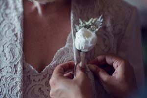 Close-up of hands pinning a white flower buttonhole onto the lace jacket of a person wearing a matching lace top, suggesting a formal or special occasion such as a wedding.