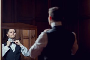 A man in formal attire adjusts his bow tie whilst looking at himself in a mirror, standing in a room with dark wooden panelling.
