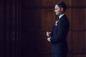 A man in a dark suit stands in profile against a wooden panelled wall, adjusting his cufflinks. He wears a buttonhole and looks thoughtful, with soft lighting highlighting his face and suit.