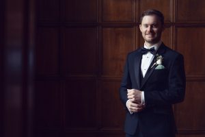 A man in a black tuxedo with a bow tie and buttonhole stands smiling in front of a wooden panelled wall, hands clasped in front of him.