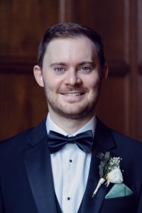 A man in a black dinner suit with a black bow tie and white dress shirt smiles at the camera. He has short brown hair, light facial hair, and a white rose buttonhole. The background is brown wood panelling.