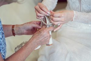 Close-up of two women’s hands as one helps the other fasten a jewelled strap on a white bridal shoe. The woman being helped is wearing a lace wedding dress and an engagement ring.