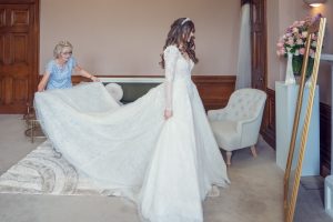 A bride in a long white wedding dress stands as an older woman arranges her train. They are in a warmly lit room with a beige armchair, large mirror, and a vase of pink flowers.