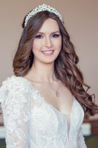 A woman with long, wavy brown hair wears a jewelled headband and a white lace wedding dress with floral details. She is smiling and looking at the camera against a softly blurred background.
