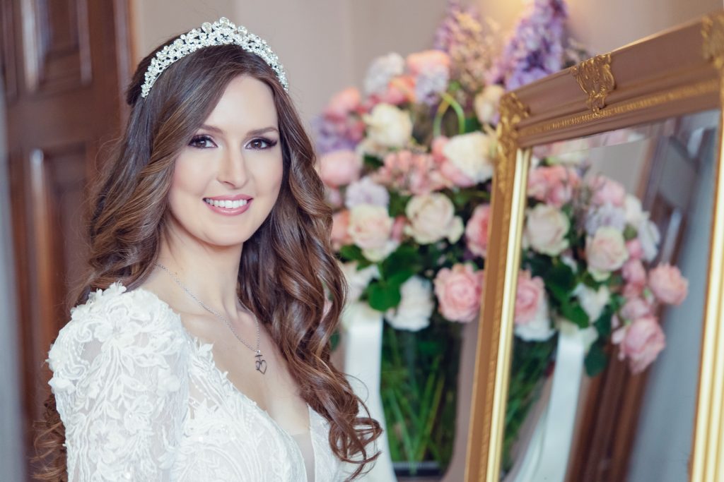A smiling bride with long, wavy brown hair and a jewelled headpiece stands next to a mirror. She wears a white lace gown and a heart-shaped necklace, with pink and white flowers in the background.
