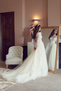 A bride in a long, elegant white wedding dress and a tiara stands in front of a large ornate mirror, admiring her reflection in a softly lit, stylish room with a white armchair and a bouquet of flowers.