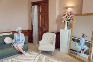 A woman in a pale blue outfit and hat sits smiling on a green sofa in an elegant room, with a large mirror reflecting her image, a vase of flowers, and a white chair nearby. A veil hangs on a wooden door.