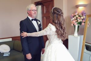 A bride in a white dress stands facing an older man in a suit, likely her father, touching his shoulders in a warmly lit room with a sofa, flowers, and a large mirror in the background.