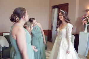 A bride in a white gown and tiara smiles at three bridesmaids in sage green dresses, who are gathered around her in a softly lit room decorated with flowers.