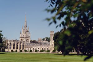 A historic, Gothic-style building with a tall clock tower and spire stands on a well-manicured lawn, partially framed by green leafy branches under a clear blue sky.