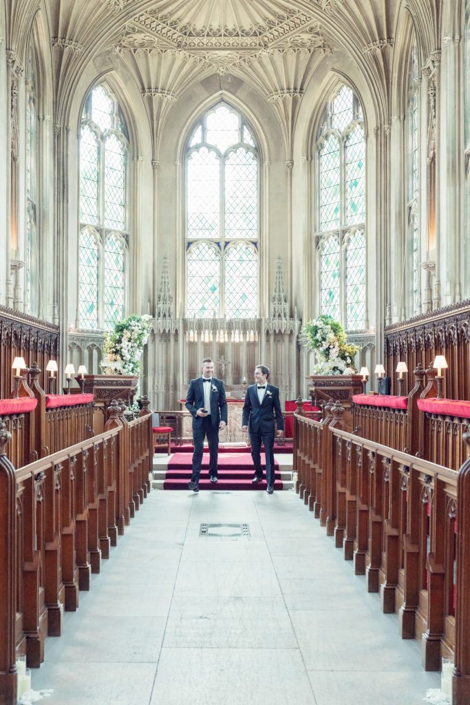 Two men in formal suits stand at the front of an ornate, empty chapel with tall arched windows, intricate stonework, wooden pews, and floral arrangements near the altar.