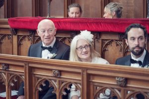 An elderly man and woman, both dressed formally, sit next to a younger man in a church pew. The woman wears a white floral headpiece. Ornate wood panelling and red velvet line the background.