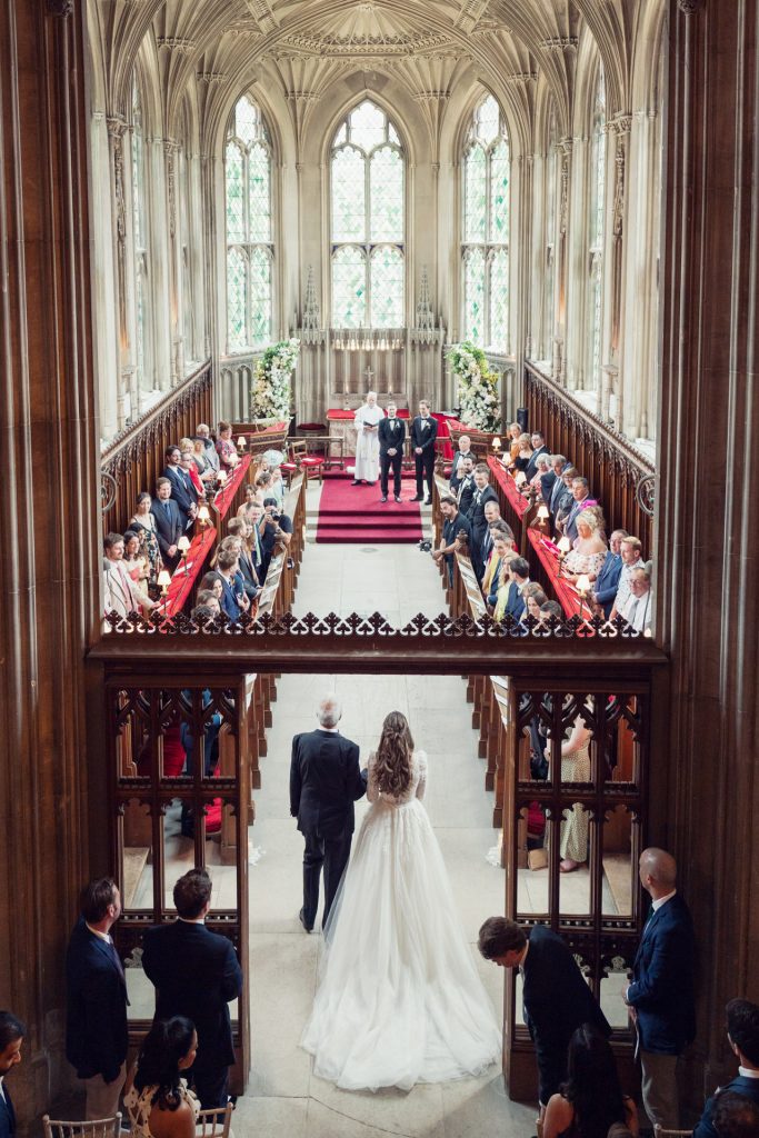 A bride in a white gown walks down the aisle with an older man in a grand, cathedral-like hall filled with seated guests, towards the altar where the groom and officiant wait. Light streams through large stained glass windows.