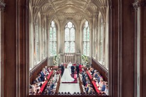 A wedding ceremony takes place inside a grand, gothic-style church with arched ceilings and large stained glass windows. Guests sit in pews, facing the bride and groom at the altar alongside officiants.