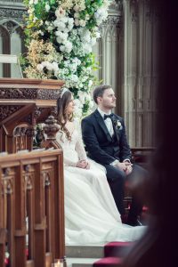 A bride and groom sit side by side in formal attire inside a church, surrounded by wooden pews and a large floral arrangement, during their wedding ceremony.