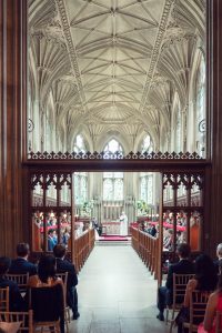 A wedding ceremony takes place in a grand, ornate church with vaulted ceilings, stained glass windows, and guests seated in wooden pews facing the altar.