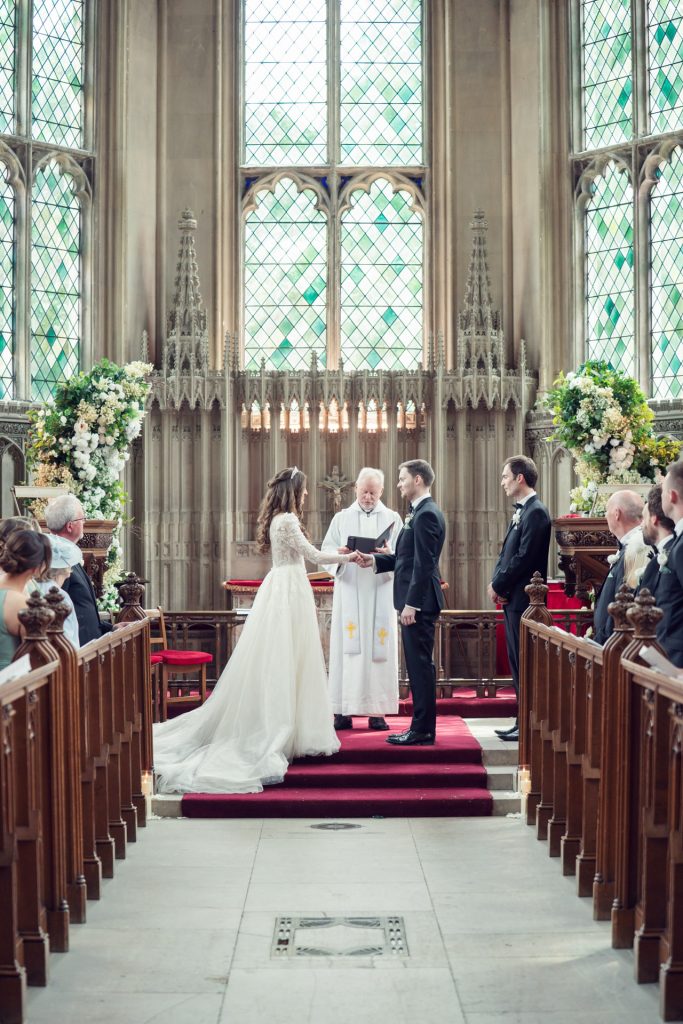 A bride and groom stand before a priest at the altar in a church, surrounded by wedding guests seated in pews. The scene is elegant, with large stained glass windows and floral arrangements decorating the altar.
