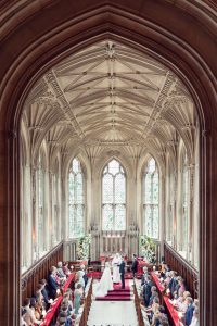 A bride and groom stand with an officiant at the altar of a grand gothic-style chapel, surrounded by guests seated in wooden pews beneath ornate vaulted ceilings and tall arched windows.