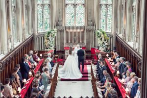 A bride and groom stand at the altar in a grand church, surrounded by guests seated in pews. The church features tall stained-glass windows, ornate woodwork, and large floral arrangements.