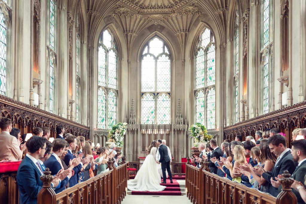 A bride and groom kiss at the altar of a grand, gothic-style church as wedding guests seated in pews applaud and celebrate. Sunlight streams through tall stained glass windows, illuminating the ornate interior.
