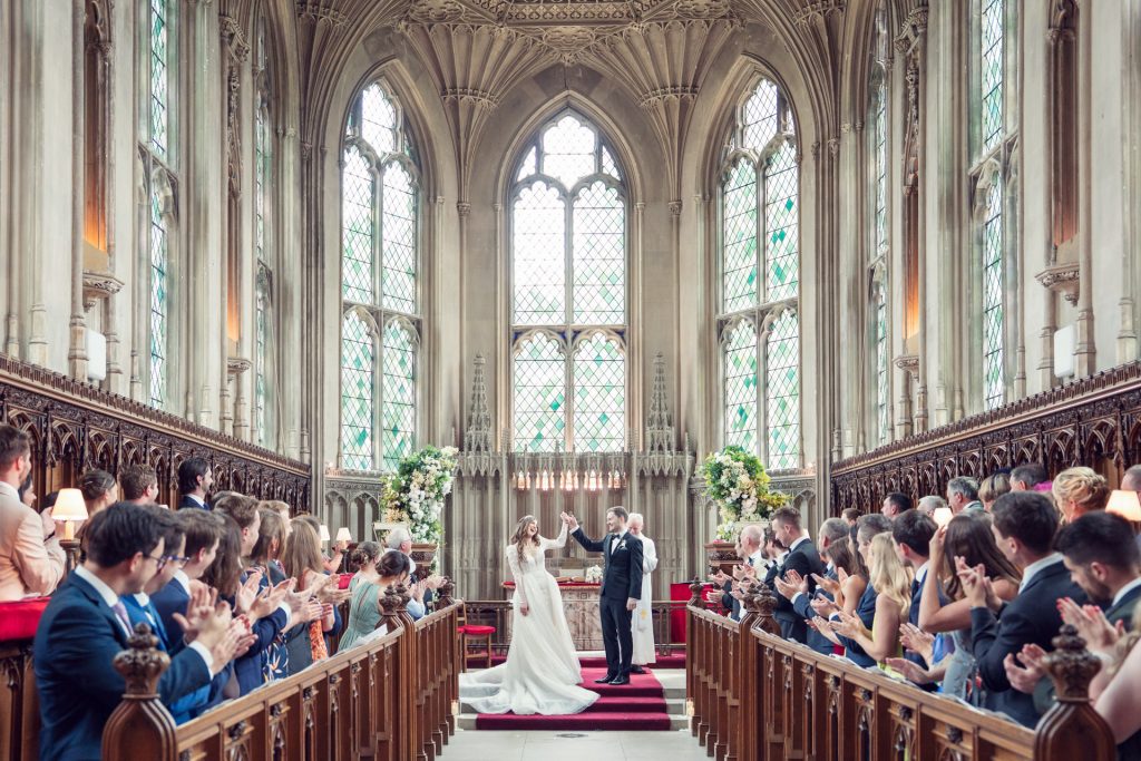 A bride and groom hold hands at the altar of a grand, gothic-style church, surrounded by applauding guests seated in pews, with tall stained glass windows in the background.