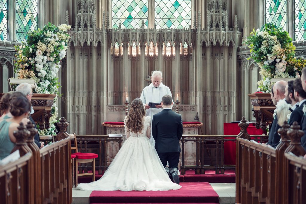 A bride and groom kneel at the altar in a church, facing a priest during their wedding ceremony, surrounded by guests seated in pews and floral arrangements on either side.