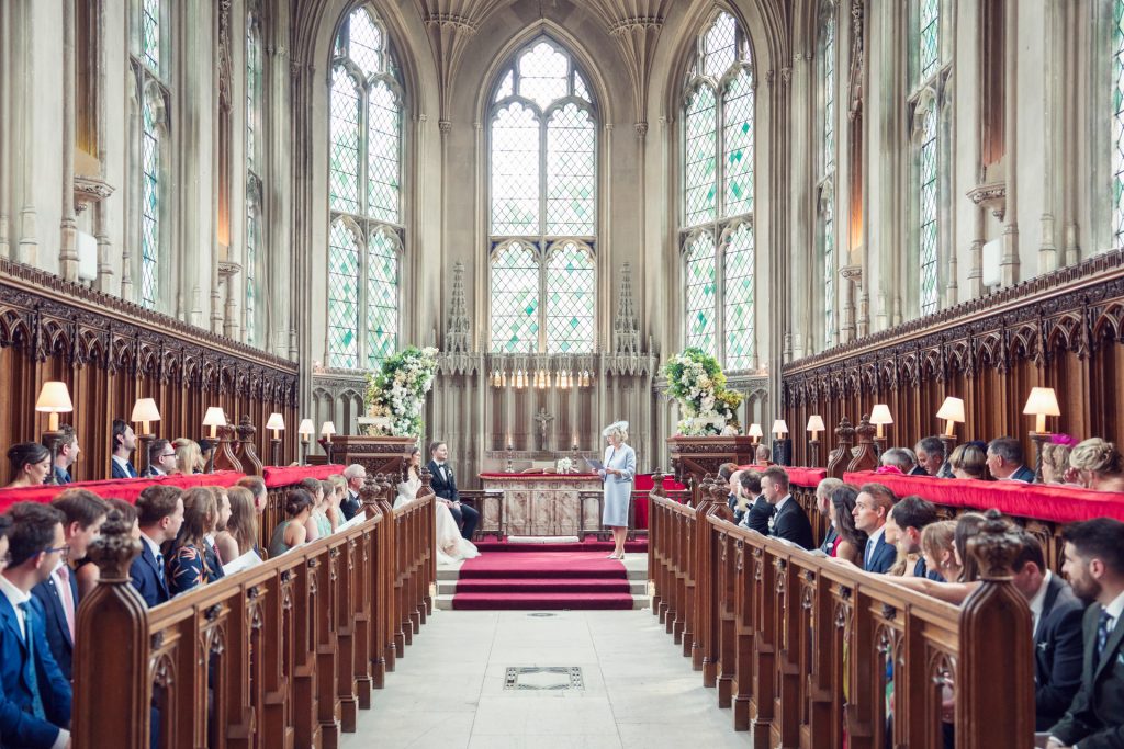 A wedding ceremony inside a grand, historic church with tall stained glass windows, ornate wooden pews, and guests seated on both sides facing the marrying couple at the altar.