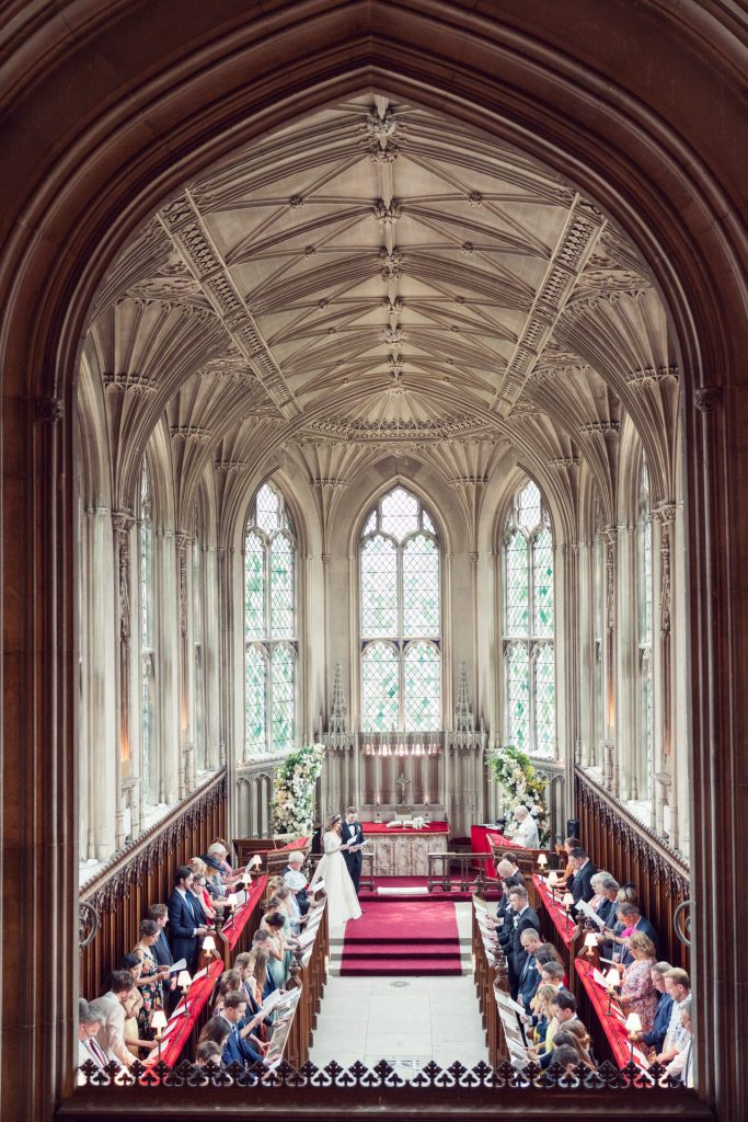 A wedding ceremony takes place in a grand, ornate hall with high vaulted ceilings, tall arched windows, and rows of seated guests on red velvet benches. The couple stand at the front, exchanging vows.