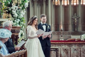 A bride and groom stand together in a church, holding papers and smiling during their wedding ceremony. Elegantly dressed guests sit nearby, and the altar is decorated with flowers and candles.