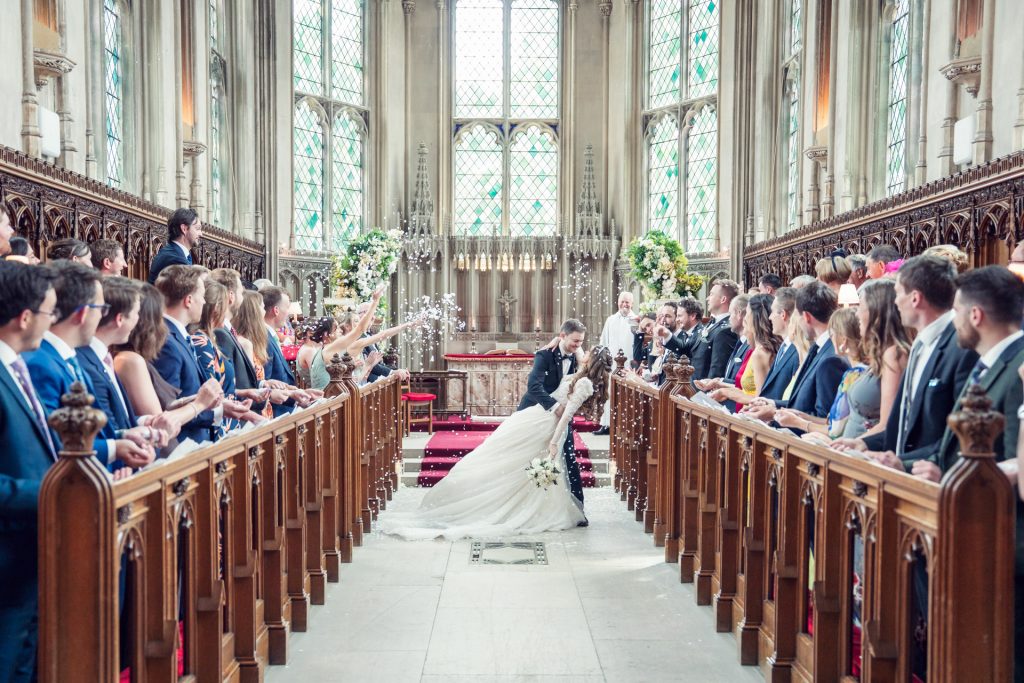 A bride and groom kiss at the altar of a church as guests seated on both sides of the aisle celebrate by throwing confetti. The church features tall stained glass windows and ornate wooden pews.