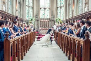 A bride and groom kiss at the altar in a grand, stained glass chapel as guests seated on both sides cheer and toss confetti. The scene is joyful and elegant, with sunlight streaming through tall windows.