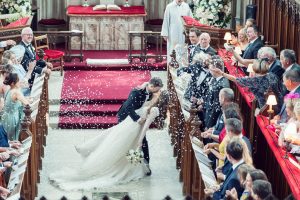 A bride and groom kiss at the altar in a church as guests shower them with white confetti. The pews are filled with family and friends, and the altar is decorated with flowers. The scene is joyful and celebratory.