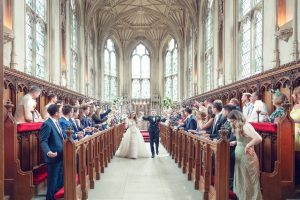A bride and groom walk down the aisle of a grand, ornate church as wedding guests on both sides celebrate and toss confetti. The couple smile, holding hands, under the high vaulted ceiling and tall stained-glass windows.