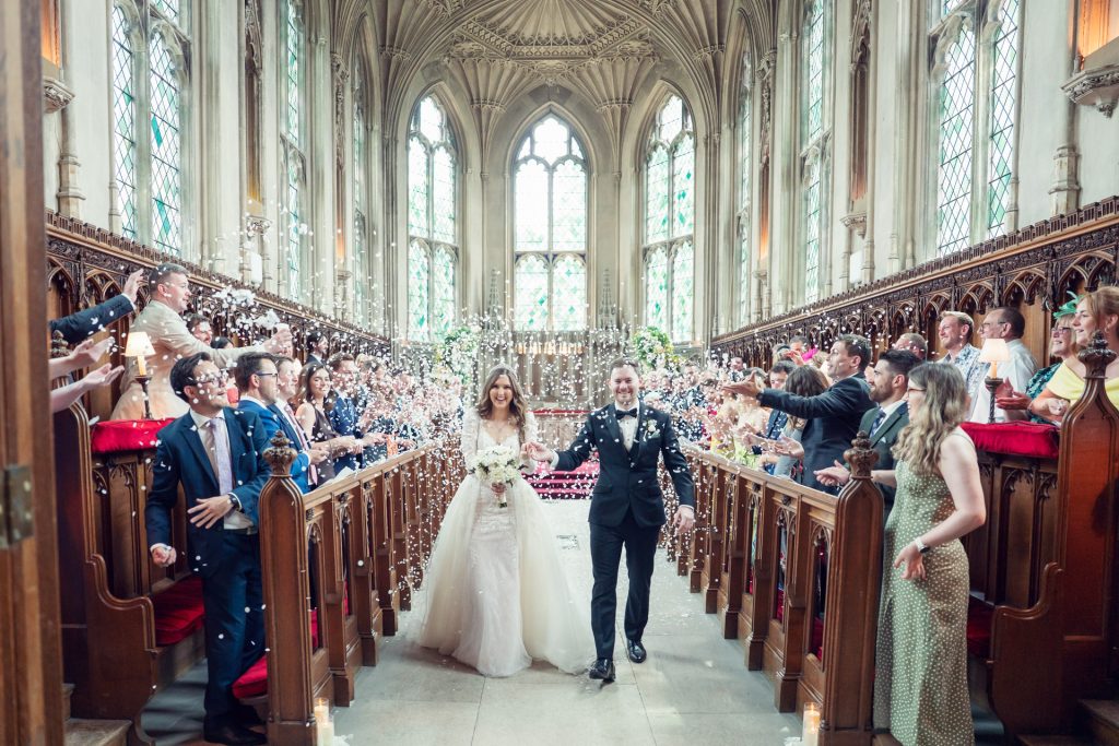 A bride and groom walk down the aisle of a grand, ornate church as guests on both sides throw confetti, celebrating their wedding. The couple smile, holding hands, surrounded by tall stained glass windows and intricate architecture.